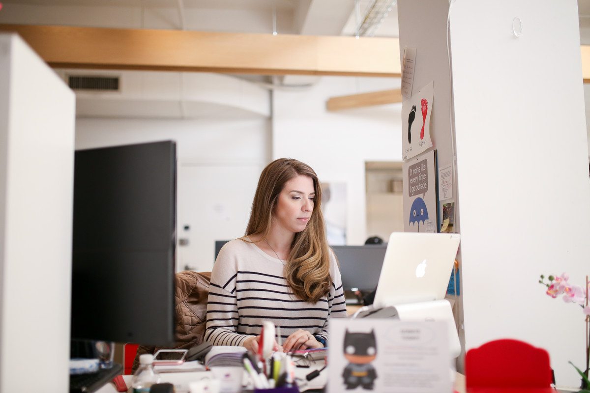 Lauren Bradley - Founder of The Officials, Lead Trainer for Executive Assistants, Typing at a Macbook at her desk. Lauren has on a white shirt with blue stripes and has long blond hair cascading over her shoulder.